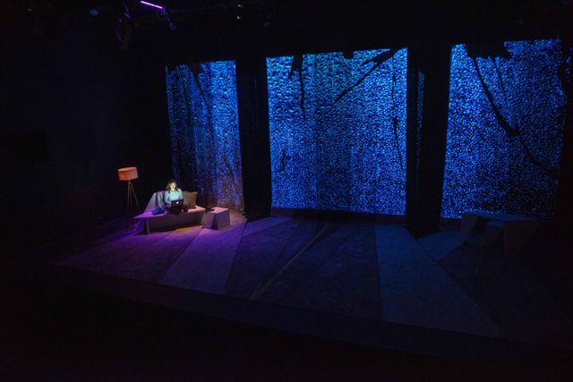High side angle of a mostly bare stage, a woman is on a couch in a pool of light and the glow of her laptop -- large screens with rain projected behind her