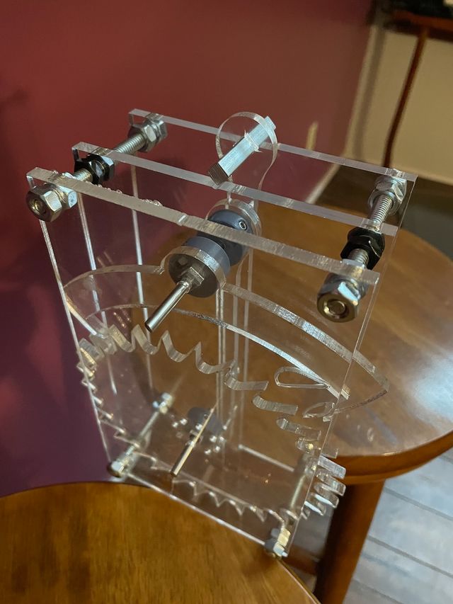 Prototype of a clear acrylic mechanical clock movement, just the pendulum and escape mechanisms, sitting across two stools so the pendulum can hang between