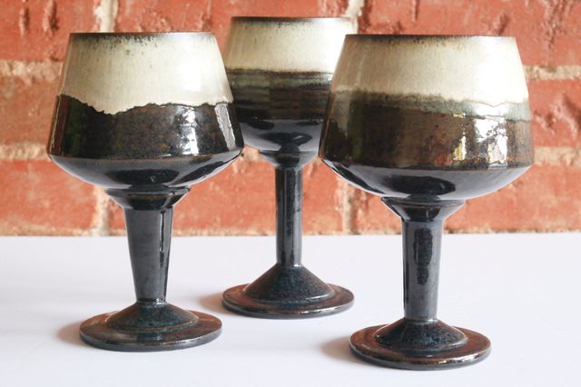 Three large wine goblets with stepped bases and slightly flared stems up to round-bottom bowls that angle in from a wide base to a narrower rim. glazed in a deep blue black from the base, turning white in the top half of the cup.